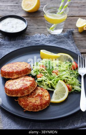 Geräucherte Lachsburger, Pasteten oder Kuchen serviert mit Krautsalat und Zitronenscheiben auf einem Teller auf rustikalem Holztisch mit Tzatziki-Sauce und Wasser, vertikal Stockfoto
