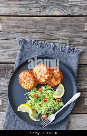 Geräucherte Lachsburger, Pasteten oder Kuchen serviert mit Krautsalat und Zitronenscheiben auf einem Teller mit Gabel auf rustikalem Holztisch, vertikale Ansicht von oben, fl Stockfoto