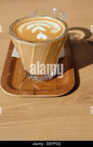 Flache weiße Nahaufnahme auf einem Holztablett, begleitet von einem Glas Wasser und einem kleinen Keks, auf einem Tisch in einem modernen Café. Stockfoto