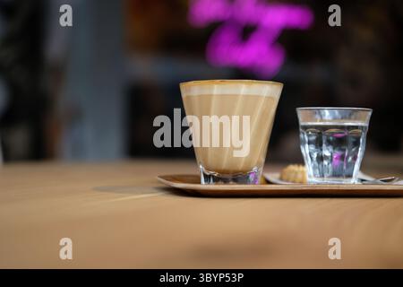 Flaches weiß mit Wasser und Keks auf Holztablett im modernen Café – gemütliche Kaffeepause, entspannender Lifestyle-Moment mit warmem Licht und weichem Bokeh. Stockfoto
