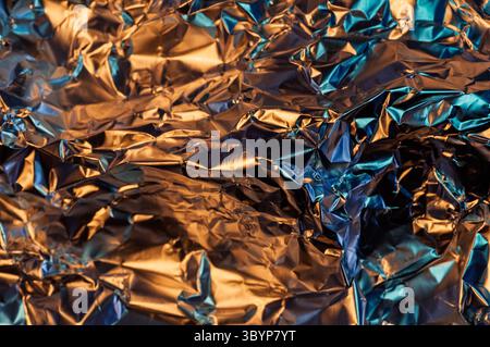 Eine Nahaufnahme aus zerknitterter Aluminiumfolie, die eine Mischung aus warmem Orange und kühlem blauem Licht reflektiert und ein strukturiertes und abstraktes Bild erzeugt.“ Stockfoto