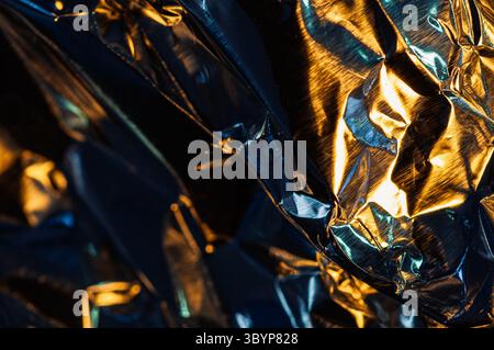 Eine abstrakte Nahaufnahme aus zerknitterter Metallfolie, die eine Mischung aus warmem goldenem und kühlem blauem Licht reflektiert. Stockfoto