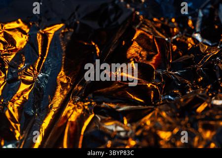 Nahaufnahme aus zerknitterter Aluminiumfolie, die warmes, goldenes Licht reflektiert. Die kontrastierenden Schatten und Lichter erzeugen eine dynamische und abstrakte Komposition. Stockfoto