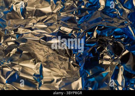 Eine Nahaufnahme aus zerknitterter Aluminiumfolie, beleuchtet mit einer Mischung aus warmem und kühlem Blau, die eine dynamische und abstrakte visuelle Textur erzeugt.“ Stockfoto