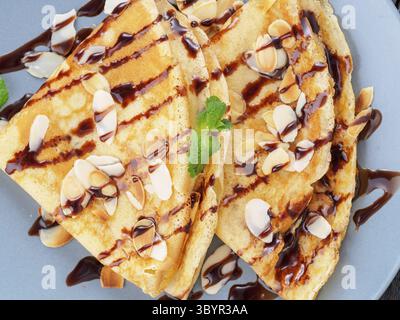 Zwei Pfannkuchen mit Schokoladensirup, Mandelflocken auf Platte, Honig fließt aus Löffel, Draufsicht, Makro Stockfoto