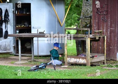 Ein Kleinkind mit blauer Kappe spielt neben einem alten Metalltisch vor einer rustikalen Werkstatt. Ein Balance Bike liegt neben ihm auf dem Boden und schafft eine nostalgische Co Stockfoto