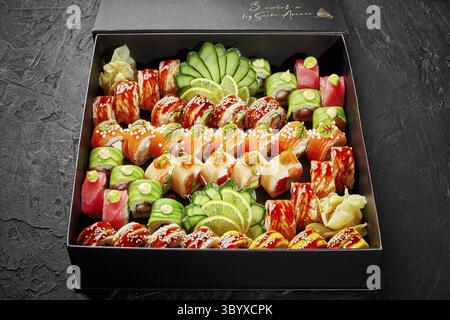 Großes Set mit verschiedenen japanischen Brötchen mit Thunfisch, Lachs und Aal, verpackt in einer Pappschachtel mit eingelegtem Ingwer, Gurkenscheiben und Zitrone, auf schwarzem Backen Stockfoto