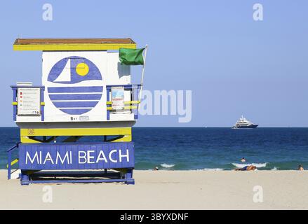 Miami Beach, USA - 9. Mai 2015: Ein Strandabschnitt mit Rettungsschwimmer und einigen Menschen, die sich sonnen oder schwimmen, eine Yacht auf dem Meer in der Bucht Stockfoto