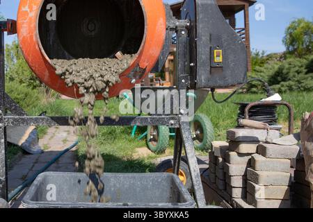 Nahaufnahme von feuchtem Betonaufwirbeln in einem orangen, tragbaren Mischer mit sichtbaren Kieselsteinen vor dem Hintergrund von Pflastern und Werkzeugen auf der Baustelle Stockfoto