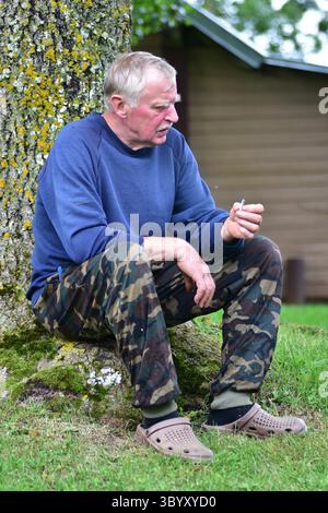 Ein älterer Mann in Tarnhose und blauem Pullover sitzt unter einem Baum auf grünem Gras, hält eine Zigarette und blickt nachdenklich in die Ferne. Rustikal Stockfoto