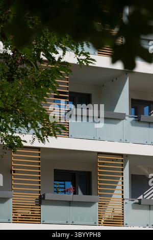Außenansicht eines modernen Apartmenthauses an einem sonnigen Tag. Die Fassade verfügt über zeitgenössische Balkone mit Milchglasgeländern und dekorativen Holzelementen Stockfoto