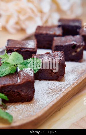 Üppige Schokoladen-Brownies werden mit frischen Minzblättern und einem Zuckerstäuben garniert, elegant auf einem Holztablett angeordnet für ein köstliches Dessert-Erlebnis Stockfoto