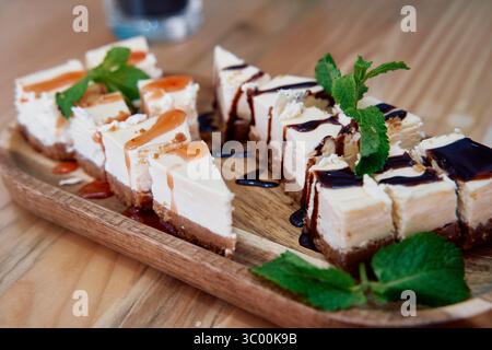 Eine Auswahl von Käsekuchen-Scheiben mit Schokolade und Karamell, geschmückt mit frischer Minze, präsentiert auf einem Holztablett. Stockfoto
