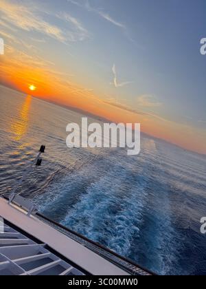 Wunderschöner Sonnenuntergang von einem Kreuzfahrtschiff aus gesehen, mit goldenen Reflexen auf ruhigem Meer und fernen Bergen am Horizont, perfekter ruhiger Reisemoment. Stockfoto