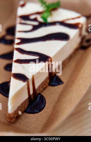 Eine üppige Käsekuchenscheibe mit Schokoladensirup liegt auf einer braunen Platte, die eine glatte Textur und eine bröckelige Kruste aufweist, perfekt zum Nachtisch Stockfoto