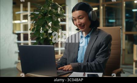 Glückliche asiatische koreanische chinesische japanische Frau hört Musik kabellos in Kopfhörern Geschäftsfrau Geschäft freudige Frau aufgeregt sorgloses Mädchen Büro Stockfoto