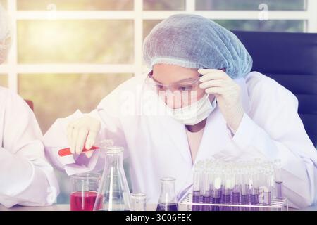 Die fokussierte Wissenschaftlerin in einem weißen Labormantel, trägt ein Haarnetz, eine Maske und eine Schutzbrille und arbeitet mit Reagenzgläsern und Bechern bei Sonnenlicht akribisch Stockfoto