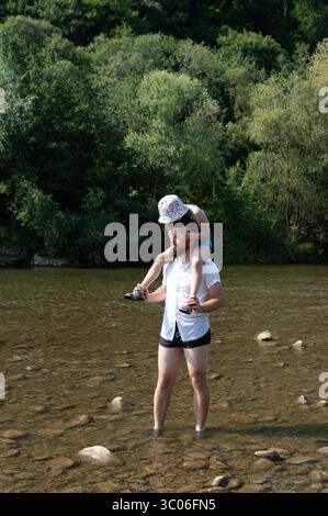 Ein Mann trägt ein Kind auf seinen Schultern durch einen flachen Fluss und genießt einen sonnigen Tag im Freien. Stockfoto