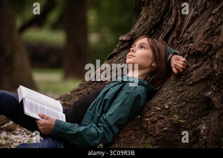 Junge, der im Park ein Buch liest, das sich an einen alten Baum lehnt. N Stockfoto