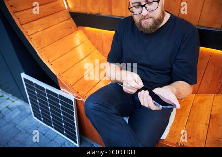 Zugeschnittene Ansicht eines Mannes, der das Smartphone mit dem Photovoltaik-Sonnenkollektor verbindet. Integration nachhaltiger erneuerbarer Energien in den Alltag, demonstrieren die praktische Nutzung von Solarstrom für Ladegeräte. Stockfoto