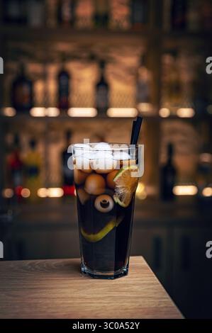 Erfrischender halloween-Cocktail mit einem gruseligen Augapfel und Eiswürfeln, serviert in einem hohen Glas an einer Bar mit verschwommenen Spirituosenflaschen im bac Stockfoto