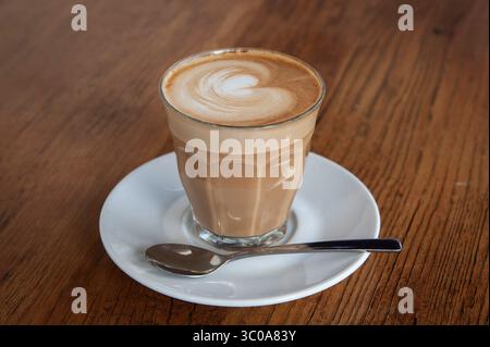 Frisch gebrühte Tasse heißer, schaumiger Cappuccino mit dekorativem Milchschaum in einem Glas auf einem Holztisch. Stockfoto