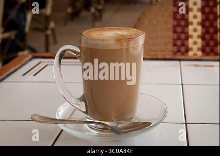 Frisch gebrühte Tasse heißen, schaumigen Cappuccino mit dekorativem Milchschaum in einem hohen Glasbecher auf einem weißen Fliesentisch. Stockfoto
