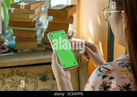 Asiatische Frau mit einer Brille, die den Finger für die Berührung am Modell des Telefons verwendet, mit leerem grünen Bildschirm mit Kopierraum in der Hand, sehen Sie sich das Smartphone im Wohnzimmer an. Stockfoto
