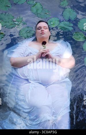 Eine große Frau schwimmt in einem weißen Kleid zwischen Seerosen, die Augen geschlossen, eine dunkle Rose in der Hand. Eine moderne, dramatische Ophelia-Szene in ruhiger, reflektierender W Stockfoto