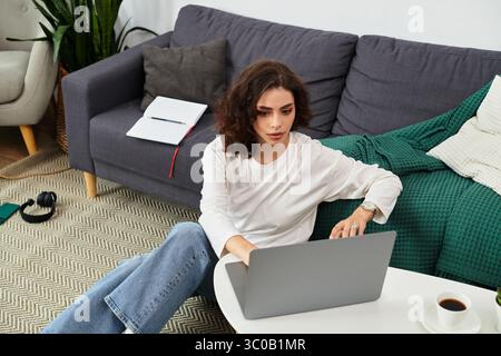 Eine junge Frau mit lockigen Haaren konzentriert sich auf ihren Laptop, während sie von ihrem komfortablen Zuhause aus arbeitet. Stockfoto