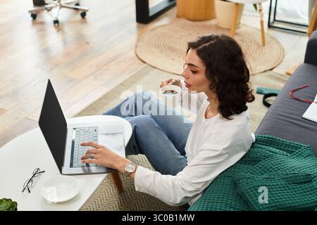 Eine junge Frau mit lockigen Haaren konzentriert sich auf ihren Laptop, während sie in einer angenehmen Umgebung Kaffee trinkt. Stockfoto
