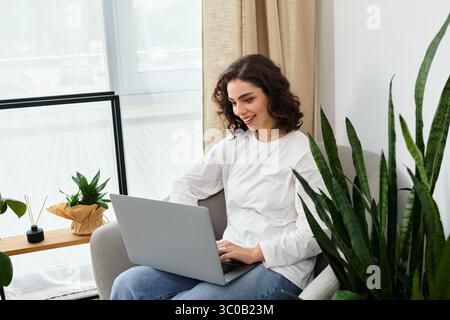Eine Frau mit lockigen Haaren konzentriert sich auf ihren Laptop in einem gemütlichen, mit Pflanzen gefüllten Arbeitsplatz zu Hause. Stockfoto