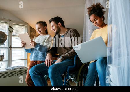 Glücklicher Vater im Rollstuhl mit interrassischen Töchtern mit Laptop zusammen, Bildung, lustiges Konzept Stockfoto