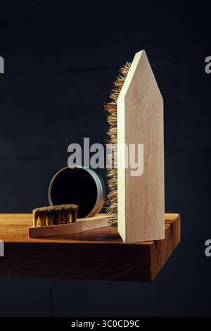Vertikales Bild mit einer Nahaufnahme traditioneller Holzschuhbürsten und einer offenen Dose dunkler schuhcreme auf einer Holzoberfläche. Eine Bürste steht aufrecht Stockfoto