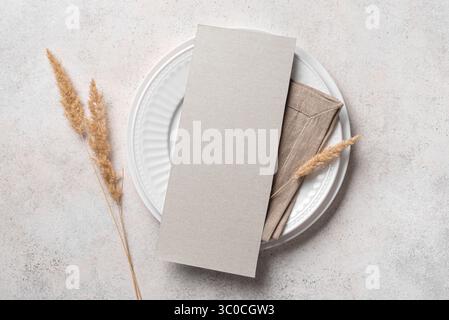 Leere 4x9 Menükarte auf weißem Teller mit Leinenserviette und trockenen Pflanzen auf neutralweißem Hintergrund, Draufsicht. Minimales elegantes Boho-Einladungskartenmockup Stockfoto