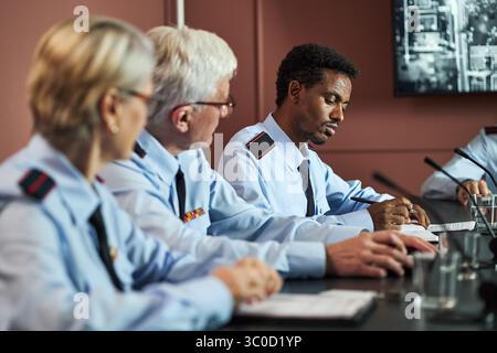 Kaukasische Frau mittleren Alters, kaukasischer Mann mittleren Alters und Schwarzer Mann mittleren Alters sitzen in Uniformen am Konferenztisch und diskutieren Dokumente während eines politischen Treffens Stockfoto