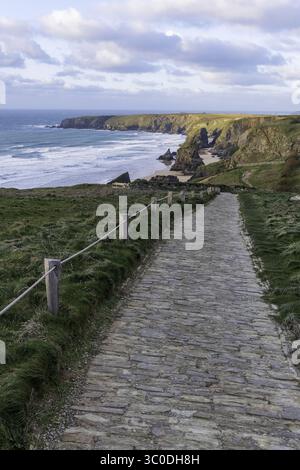 Blick auf einen Steinweg, der zur zerklüfteten Küste mit dramatischen Klippen und stürzenden Wellen unter einem ruhigen Himmel führt, Wadebridge, England, Vereinigtes Königreich Stockfoto