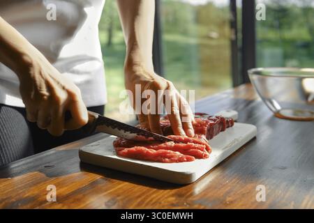 Eine Person schneidet gekonnt mageres Rindfleisch auf einem Schneidebrett in einer hellen Küche. Natürliches Licht filtert durch große Fenster und hebt das Frische hervor Stockfoto
