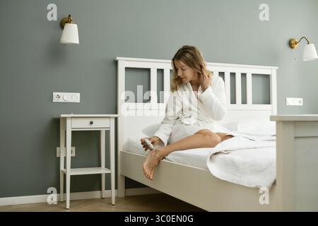 Eine Frau in einem weißen Gewand sitzt am Rand eines Bettes in einem sanft beleuchteten Schlafzimmer. Sie benutzt eine elektrische Peelingmaschine, um tote Haut von ihren Füßen zu entfernen Stockfoto