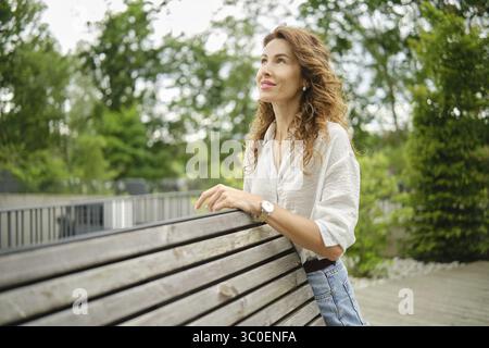 Eine Frau lehnt sich an eine Holzbank in einem üppig grünen Park und blickt nachdenklich in die Ferne. Die Szene fängt die Ruhe eines sonnigen Aftern ein Stockfoto