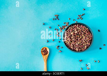 Flacher Lagen einer schwarzen Keramikschale, gefüllt mit duftenden Sichuan-Pfefferkörnern, verstreuten Gewürzen und einem kleinen Holzlöffel auf geräumigem türkisfarbenem Hintergrund Stockfoto