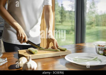Unerkennbare Frau schneidet gekonnt grünen Lauch auf einem hölzernen Schneidebrett in einer hellen Küche mit Blick auf den Außenbereich Stockfoto