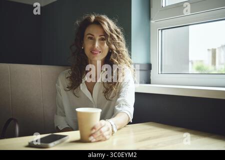 Eine Frau mit lockigen Haaren sitzt an einem Tisch in der Nähe eines Fensters und hält eine Tasse Kaffee. Sie trägt ein weißes Hemd und lächelt sanft, mit ihrem Handy neben sich. Stockfoto