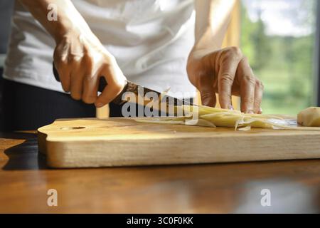 Unerkennbare Person schneidet gekonnt frischen Ingwer auf einem hölzernen Schneidebrett in einer gut beleuchteten Küche Stockfoto