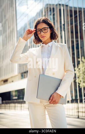 Selbstbewusste Geschäftsfrau im Freien, die einen Laptop hält, einen stilvollen Anzug trägt, in einem urbanen Stadtbild steht und Professionalität widerspiegelt Stockfoto