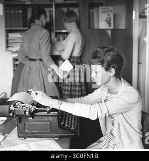 Büromädchen in den 1950ern Eine Frau sitzt vor ihrer Schreibmaschine und sieht den Text auf dem Blatt Papier in der Maschine an. Sie ist an ihrem ersten Tag in der neuen Stelle und einige der Mitarbeiter stehen im Hintergrund und sprechen, was darauf hindeutet, dass sie nicht dazugehört und es eine unsichtbare Barriere bei ihren Kollegen gibt. 1957 Stockfoto
