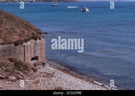 Ruhige Küstenlandschaft mit einem verwitterten Bunker an einem felsigen Ufer mit Blick auf eine ruhige Bucht mit Segelbooten, die das ruhige Wasser genießen Stockfoto