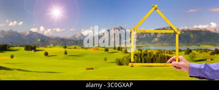 Wohnen auf dem Land, Handschuh in der Natur ein Meterstab ist gefaltet wie ein Haus und wird mit der Hand vor einer unberührte Landschaft, Handschuh in der N Stockfoto