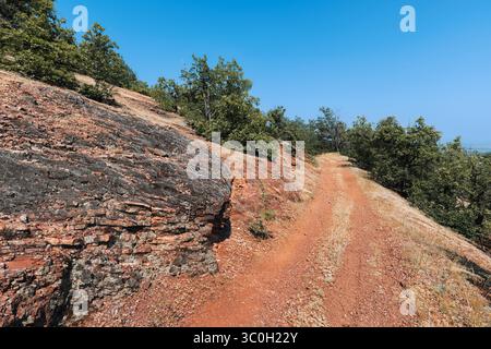 Rote Feldstraße schlängelt sich durch eine felsige Landschaft mit Bäumen und Vegetation unter einem klaren blauen Himmel und schafft eine malerische Naturszene Stockfoto