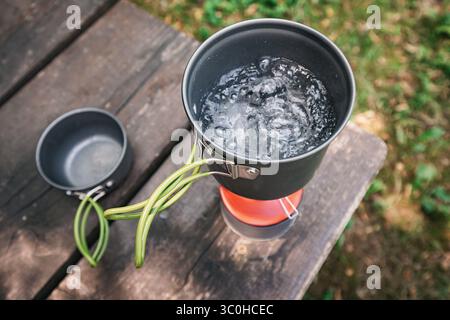 Kochendes Wasser in einem Campingtopf auf einem tragbaren Gasherd auf einem Holztisch während eines Trekkingtages Stockfoto